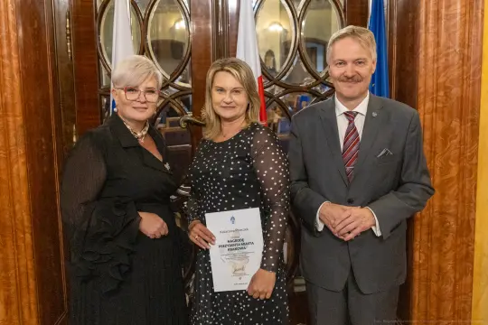 Trzy osoby w eleganckich strojach, trzymające dokumenty, stoją przed ozdobnymi drzwiami.