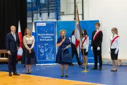 Uroczystość z przemówieniem w sali, uczestnicy w strojach galowych, flagi i baner Wydziału Edukacji.