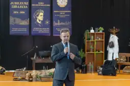Mężczyzna w garniturze przemawia do zgromadzonej publiczności w sali z dekoracjami jubileuszowymi.