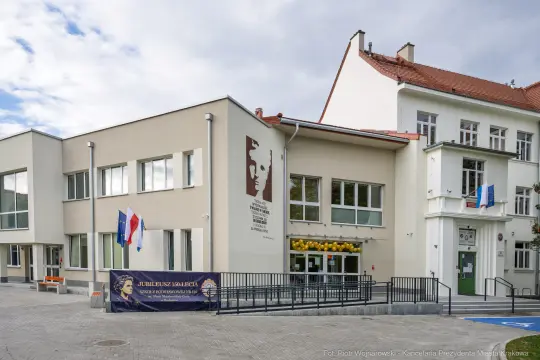 Budynek szkoły z flagami i tablicą pamiątkową na elewacji.
