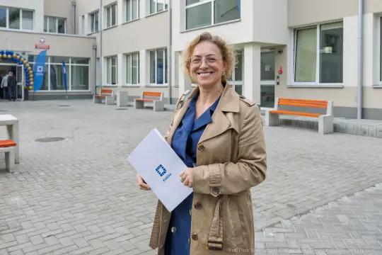 Kobieta w beżowym płaszczu trzyma dokumenty, uśmiechając się na tle budynku.