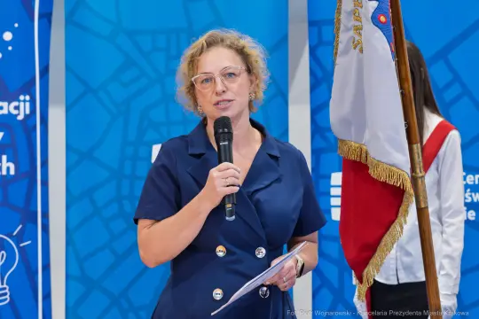 Kobieta z mikrofonem wygłasza przemówienie, obok stoi osoba z flagą.