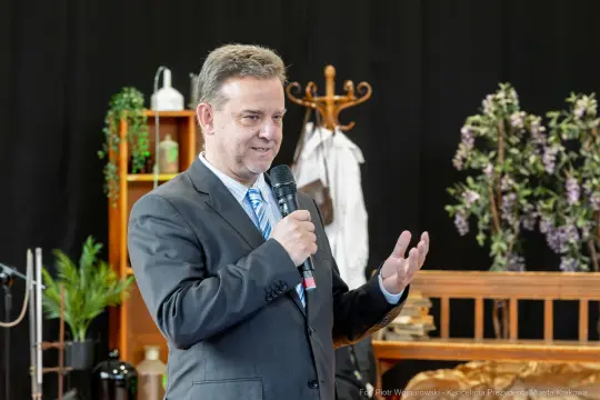 Mężczyzna w garniturze przemawia do publiczności, trzymając mikrofon. W tle dekoracje i rośliny.