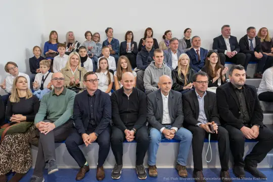 Grupa ludzi siedzących na trybunach, wśród nich dzieci i dorośli, w różnych strojach.