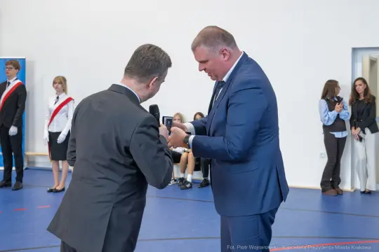 Dwaj mężczyźni w garniturach podczas uroczystości wręczenia nagrody. W tle uczniowie i nauczyciele.