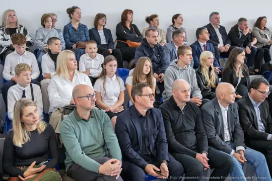 Grupa ludzi siedzących na widowni, w różnym wieku, w formalnych strojach.