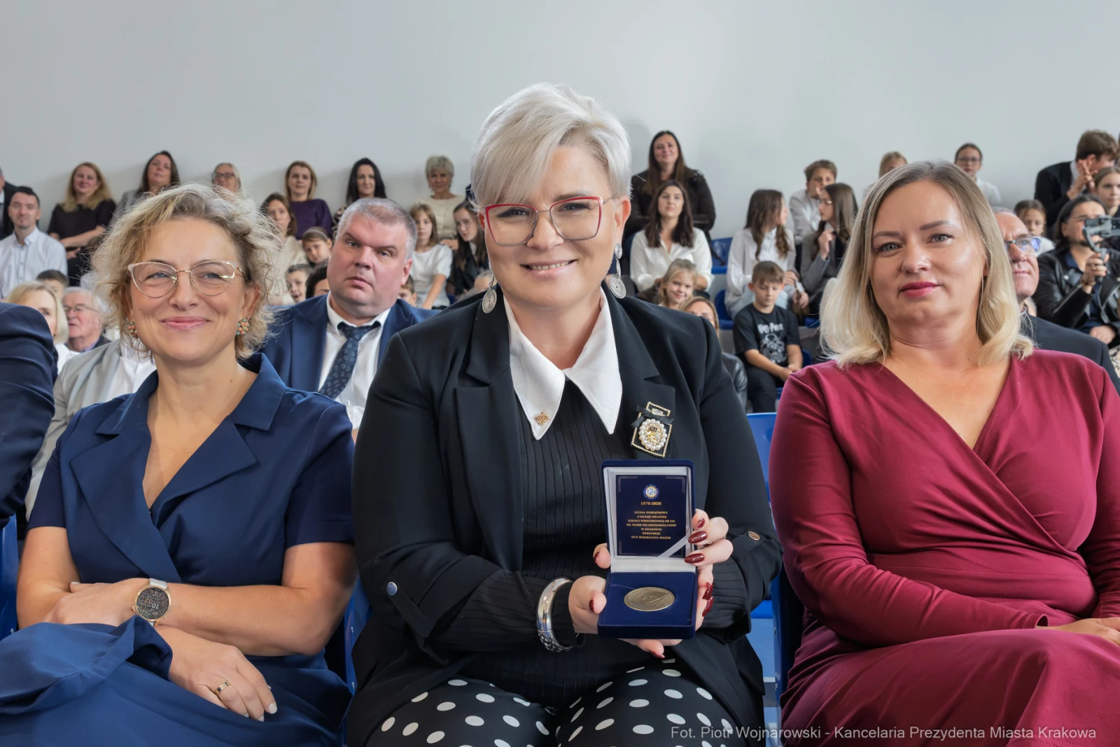 Kobieta w okularach trzyma medal, obok dwie inne kobiety w eleganckich strojach. W tle widownia.  Autor: Piotr Wojnarowski