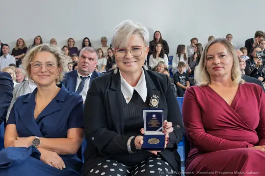 Kobieta w okularach trzyma medal, obok dwie inne kobiety w eleganckich strojach. W tle widownia.