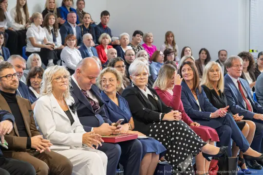 Grupa ludzi siedzących na widowni podczas wydarzenia, wśród nich kilka kobiet w eleganckich strojach.