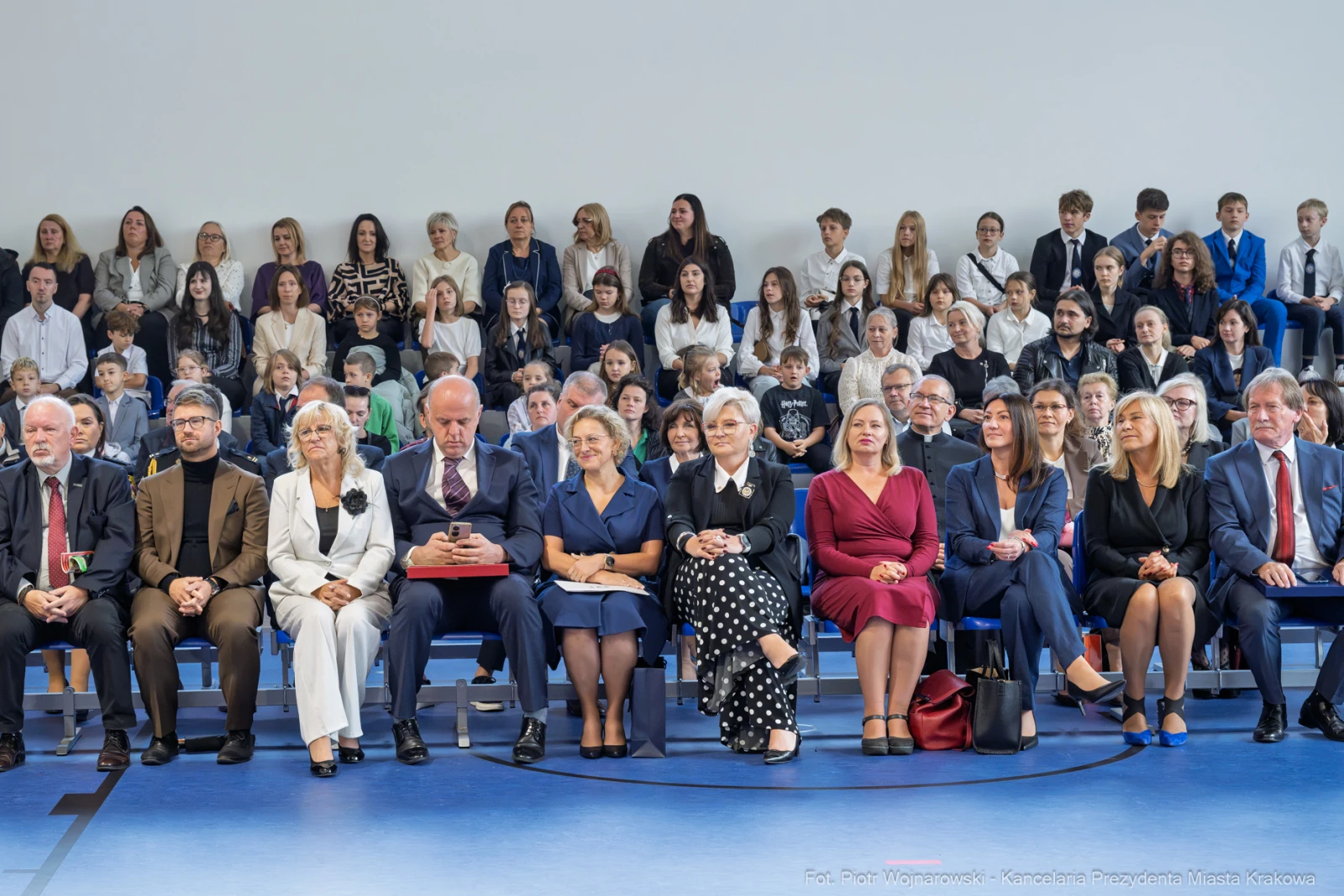 Grupa osób siedzących na widowni podczas uroczystości, w tle dzieci i młodzież.  Autor: Piotr Wojnarowski