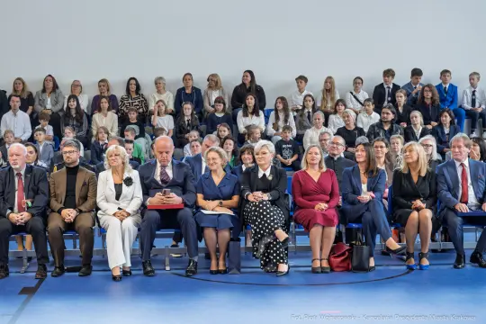Grupa osób siedzących na widowni podczas uroczystości, w tle dzieci i młodzież.