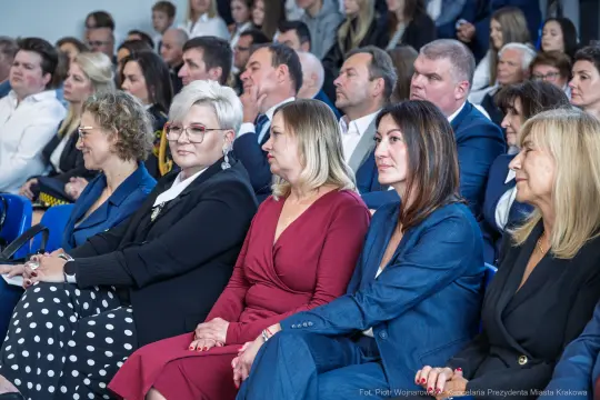Grupa osób siedzących na wydarzeniu, zróżnicowane reakcje i styl ubioru.