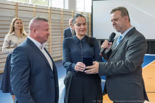 Wręczenie odznaczenia w sali sportowej, trzech osób w formalnych strojach.