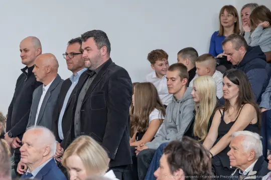 Grupa ludzi siedzących i stojących na wydarzeniu, różne emocje i interakcje.