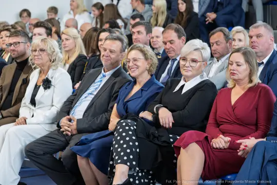Grupa osób siedzących na wydarzeniu, w różnorodnych strojach, z uśmiechami i poważnymi minami.