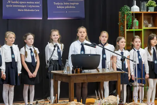 Dziewczynki w białych koszulach i czarnych spódnicach śpiewają na scenie.