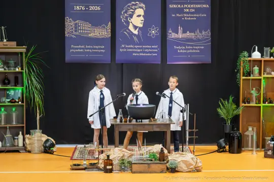 Uczniowie w białych fartuchach prezentują eksperymenty naukowe na scenie.