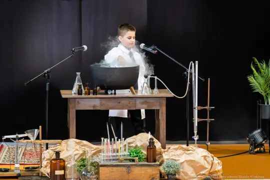 Chłopiec w białym fartuchu przeprowadza eksperyment chemiczny przy stole z przyrządami.