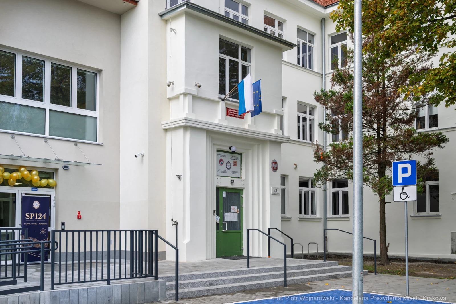 Budynek szkoły z flagą i oznaczeniem miejsca parkingowego dla osób niepełnosprawnych.  Autor: Piotr Wojnarowski