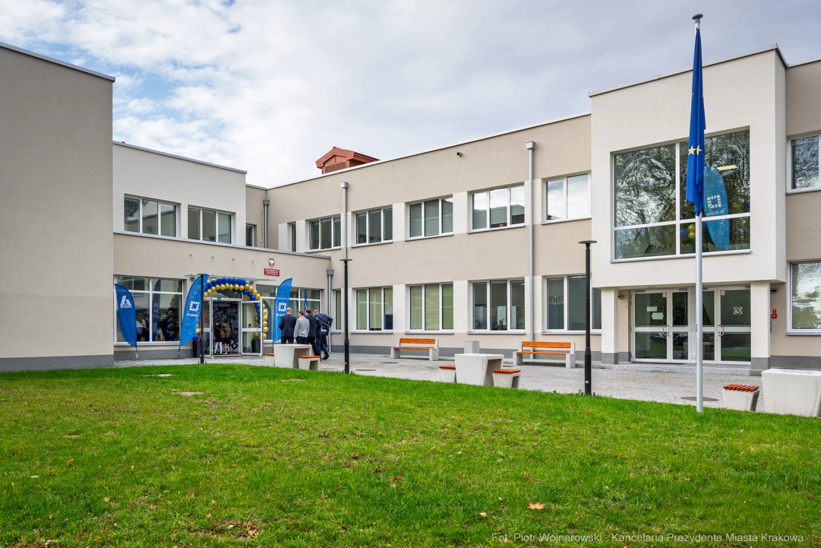Budynek szkoły z flagami, zielonym trawnikiem i osobami wchodzącymi do środka.  Autor: Piotr Wojnarowski