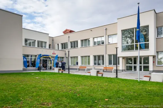 Budynek szkoły z flagami, zielonym trawnikiem i osobami wchodzącymi do środka.
