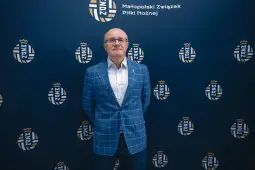 Mężczyzna w niebieskim garniturze stoi przed tłem z logo Małopolskiego Związku Piłki Nożnej.