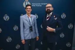 Dwaj mężczyźni w eleganckich strojach, stojący obok siebie na tle logo Małopolskiego Związku Piłki Nożnej.