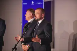 Mężczyzna w garniturze przemawia do mikrofonu, w tle inny mężczyzna.