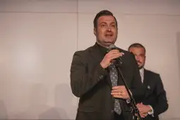 Mężczyzna w garniturze przemawia do publiczności, w tle inny mężczyzna.