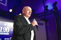 Mężczyzna z mikrofonem na scenie, w tle oświetlenie i ekran z napisem 120.