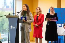 Kobieta przemawia z mikrofonem, obok dwie inne kobiety, w tle widoczna sala konferencyjna.