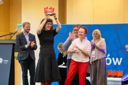 Kobieta trzymająca trofeum, otoczona przez uśmiechnięte osoby, na tle kolorowej ścianki.