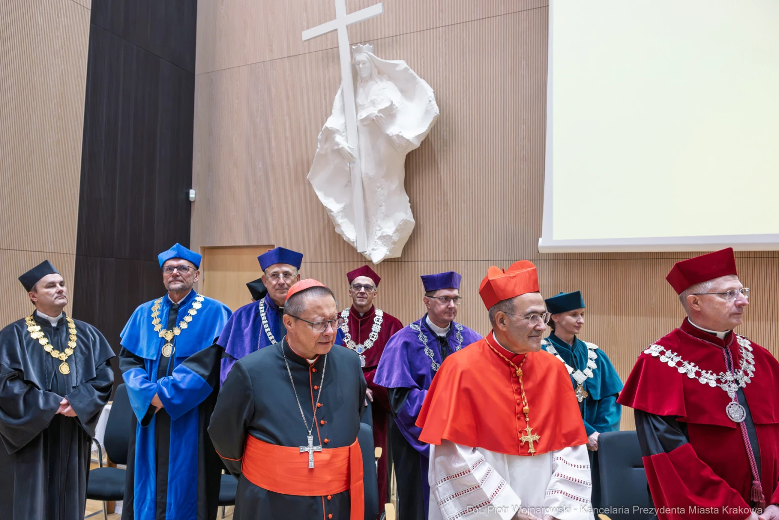 Grupa dostojników w akademickich togach, w tym kardynał, na tle nowoczesnej dekoracji.  Autor: Piotr Wojnarowski