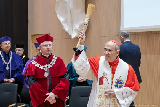 Uroczystość akademicka z osobą w szatach liturgicznych trzymającą miotłę.