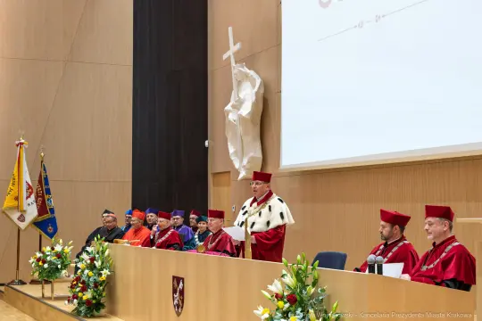 Uroczystość akademicka z mówcą i uczestnikami w togach, dekoracje kwiatowe na stole.