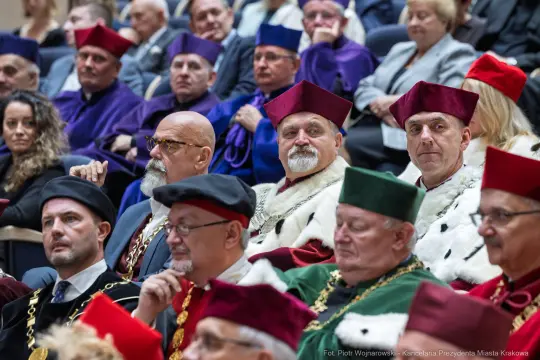 Grupa osób w akademickich togach i biretach, siedzących na uroczystości.