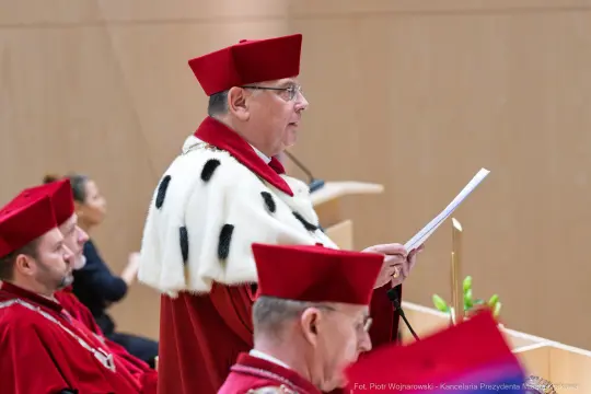 Mężczyzna w czerwonym stroju akademickim wygłasza przemówienie.