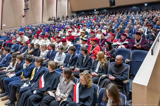 Uroczystość akademicka z uczestnikami w togach i garniturach, siedzącymi w sali.