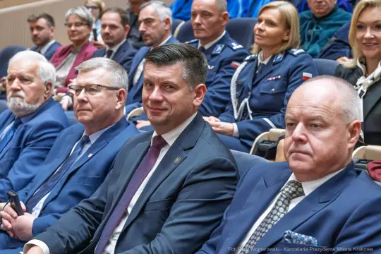 Grupa ludzi siedzących na sali, w tym mężczyzna w garniturze z uśmiechem.