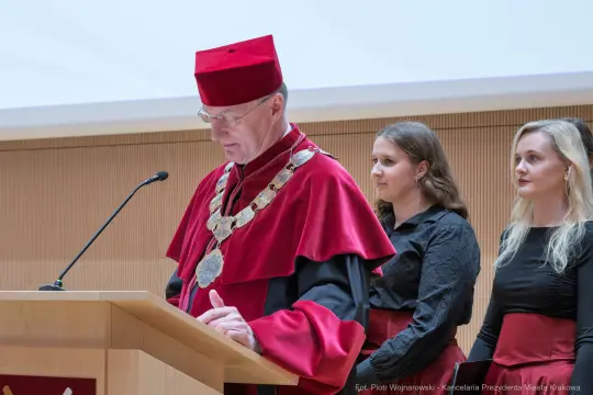 Mężczyzna w akademickim stroju wygłasza przemówienie, obok dwie kobiety w eleganckich strojach.