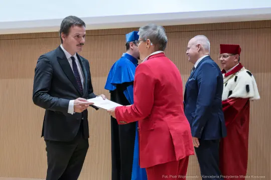 Wręczenie dyplomu przez kobietę w czerwonym garniturze mężczyźnie w garniturze. W tle inni uczestnicy ceremonii.