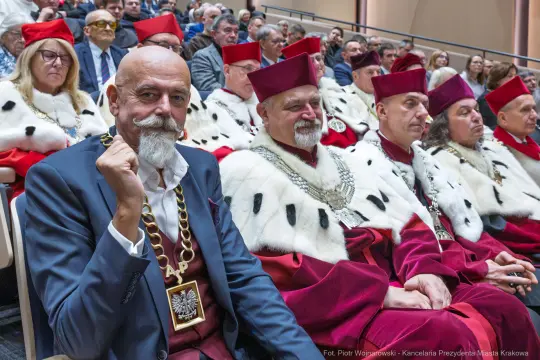 Mężczyźni w eleganckich strojach z łańcuchami i pelerynami, siedzący na uroczystości.