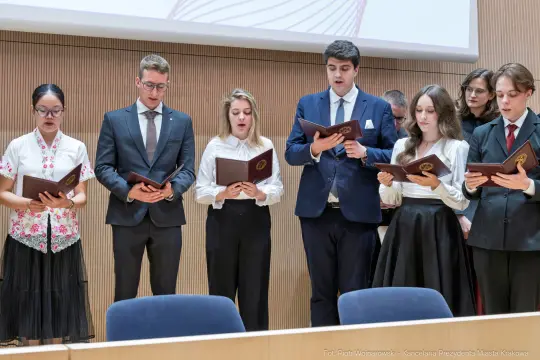 Grupa osób w eleganckich strojach śpiewa, trzymając dokumenty w rękach.
