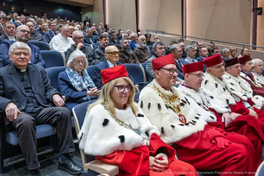 Grupa osób w ceremonialnych strojach siedzących na widowni, w tle zgromadzenie publiczności.