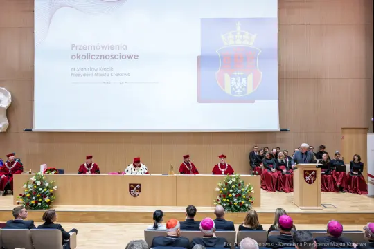 Uroczystość z przemówieniami w sali z dekoracjami i przedstawicielami władz.