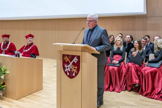 Mężczyzna przemawia na uroczystości akademickiej, w tle siedzą goście w togach.