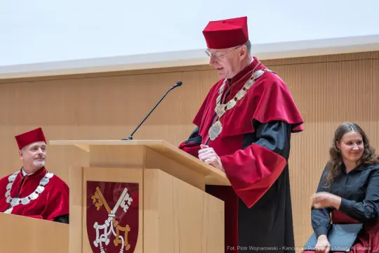 Mężczyzna w czerwonej todze i birecie przemawia do zgromadzenia. Obok siedzą inni członkowie akademickiego grona.