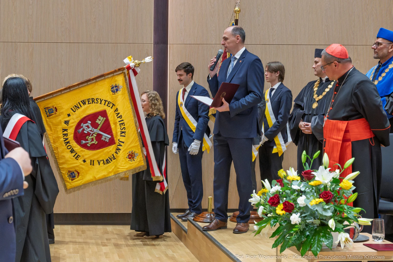 Uroczystość akademicka z flagą Uniwersytetu Jagiellońskiego i mówcą na podium.  Autor: Piotr Wojnarowski