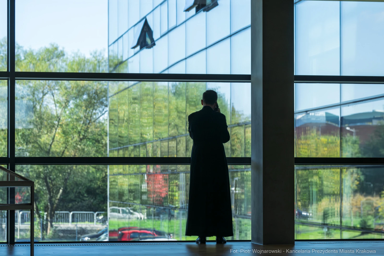 Mężczyzna w długim płaszczu stoi przy dużym oknie, patrząc na zewnętrzny krajobraz.  Autor: Piotr Wojnarowski