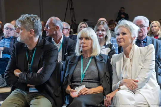 Grupa osób siedzących na konferencji, zróżnicowane reakcje i styl ubioru.